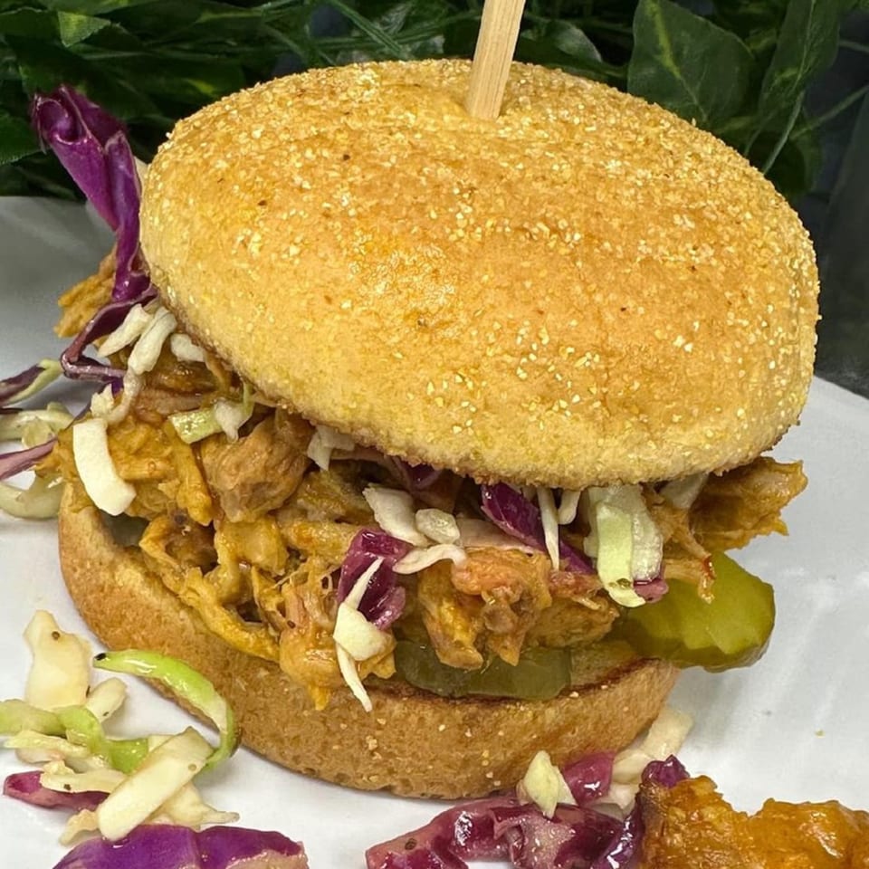 Pulled pork sandwich with coleslaw and pickles on a toasted bun, served on a white plate.