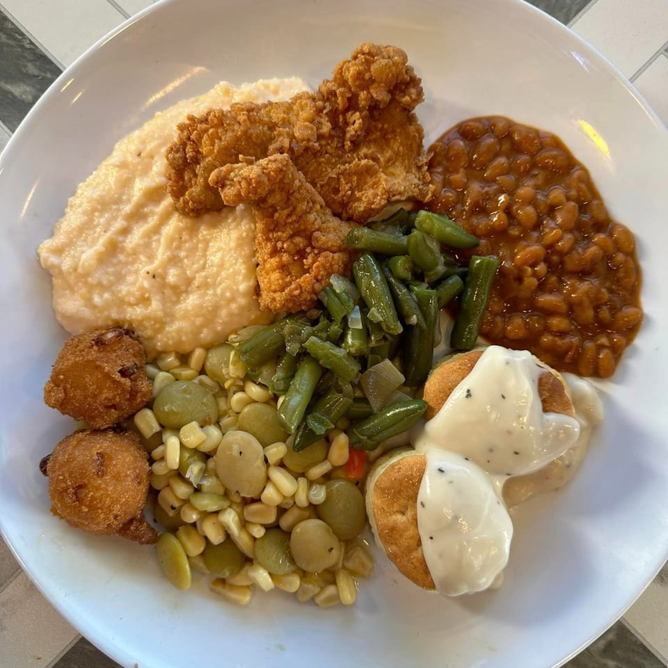A Georgia Comfort Kitchen plate with fried chicken, grits, baked beans, green beans, corn, lima beans, hush puppies, and biscuits with gravy.