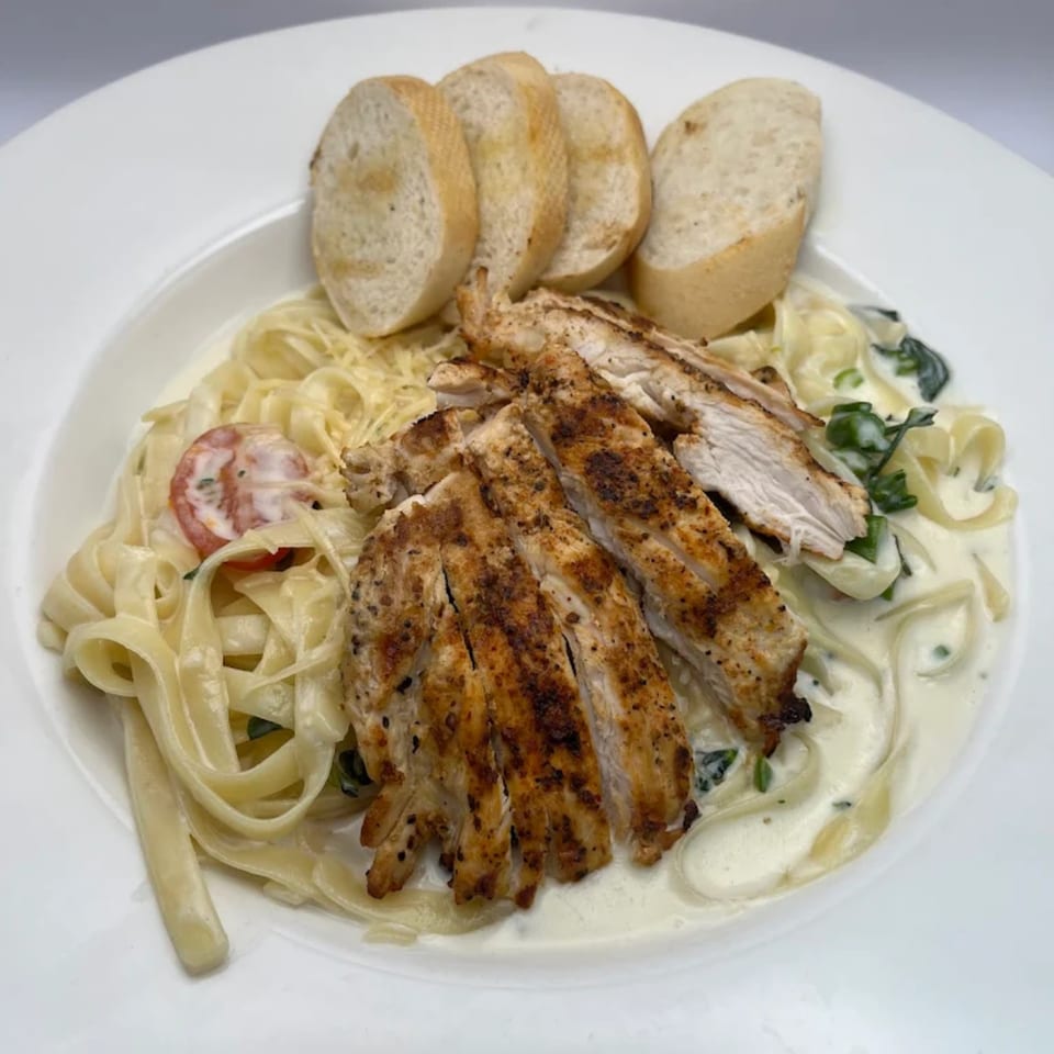 Grilled chicken fettuccine alfredo with cherry tomatoes, spinach, and garlic bread on a white plate.