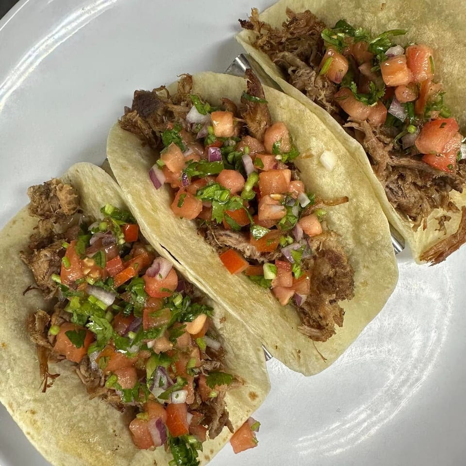 Three Georgia Comfort Kitchen tacos with shredded meat, diced tomatoes, and cilantro on a white plate.