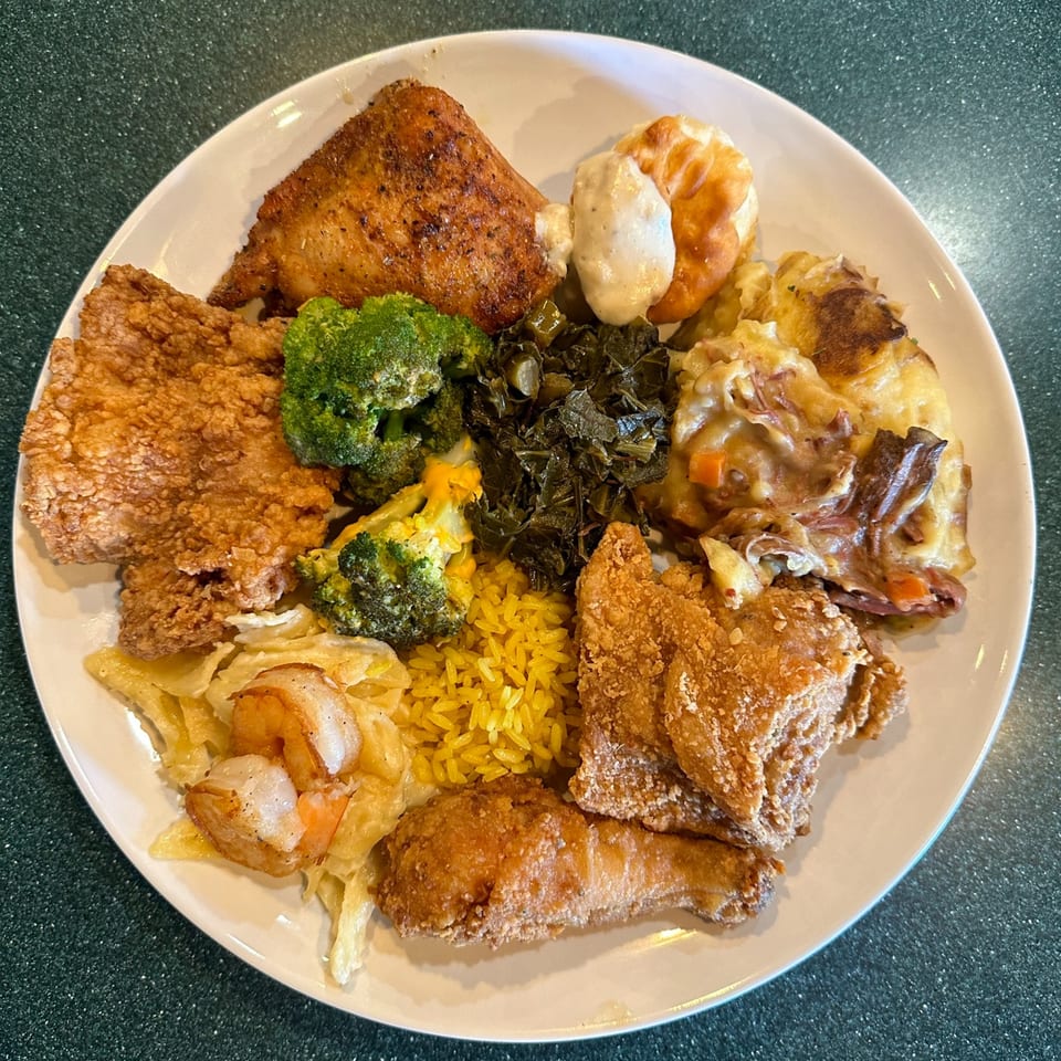 A white plate overflowing with Georgia Comfort Kitchen's fried chicken, shrimp, rice, broccoli, and collard greens.