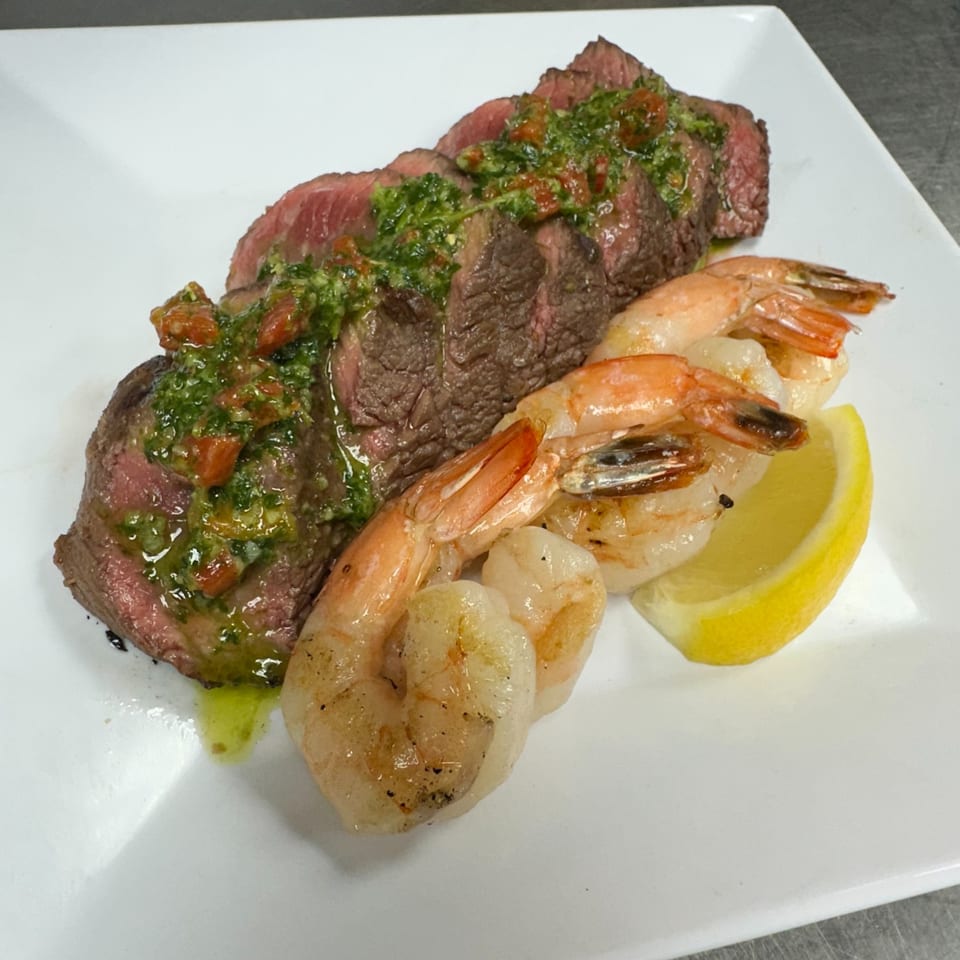 Sliced steak topped with green sauce and diced tomatoes, served with shrimp and a lemon wedge.